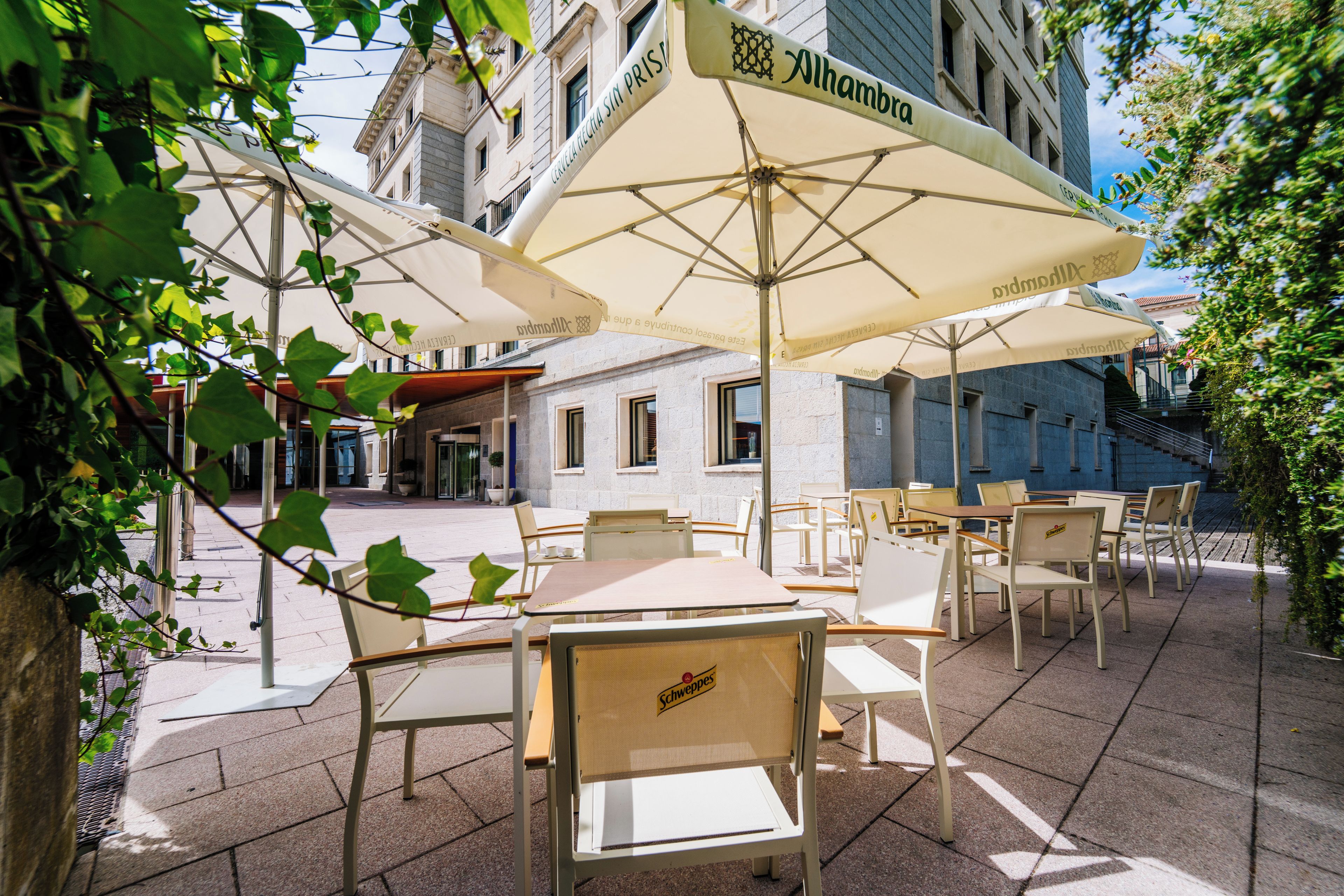 An outdoor café area with tables and chairs under umbrellas, a building in the background, and a clear sky.