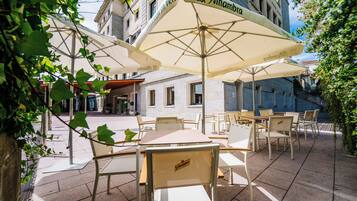 An outdoor café area with tables and chairs under umbrellas, a building in the background, and a clear sky.