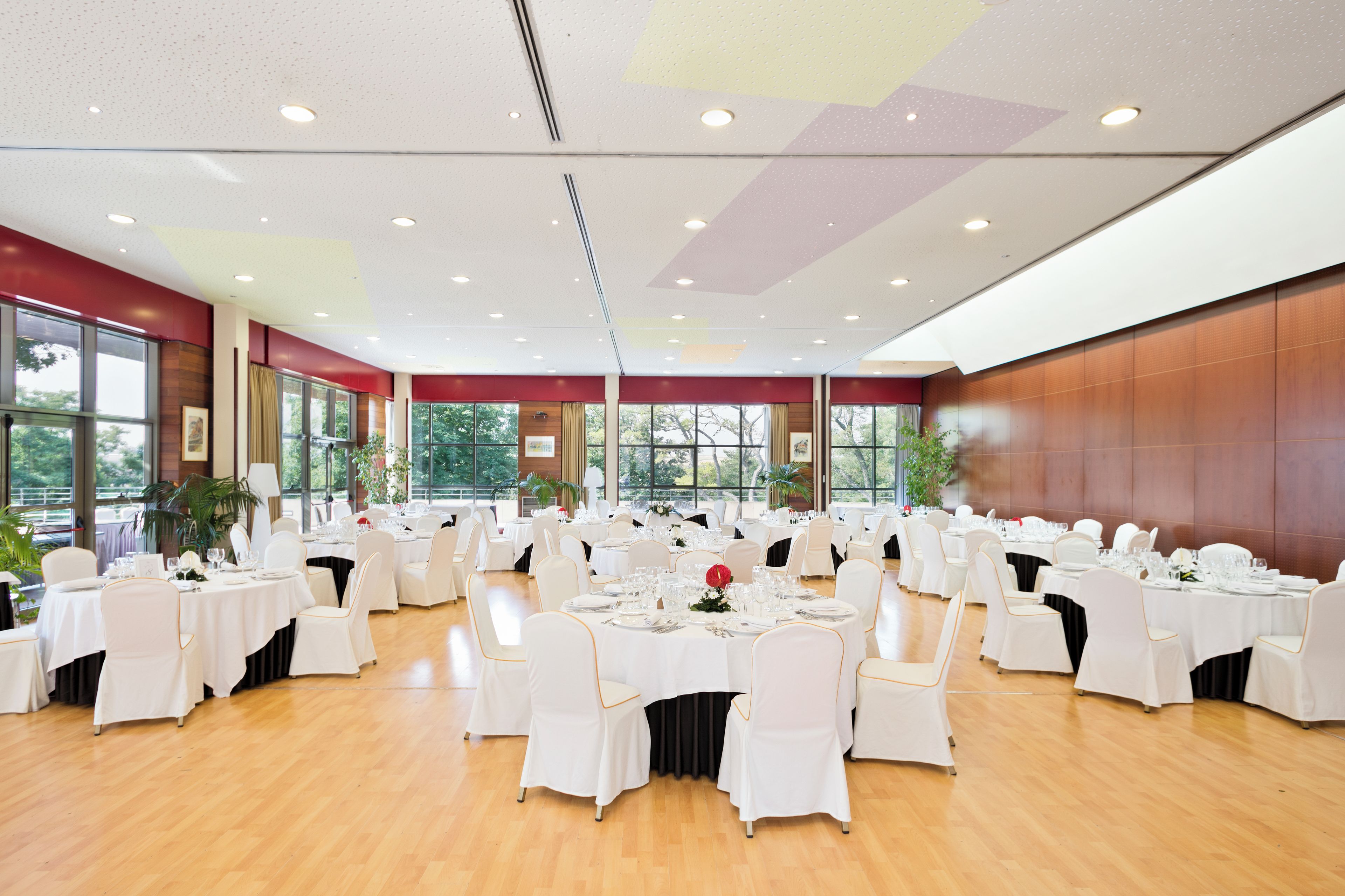 Une vaste salle de banquet avec de nombreuses tables rondes dressées pour le repas, des chaises blanches et un plafond haut doté d’un éclairage encastré.