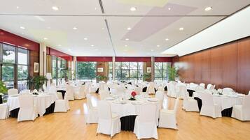 Une vaste salle de banquet avec de nombreuses tables rondes dressées pour le repas, des chaises blanches et un plafond haut doté d’un éclairage encastré.