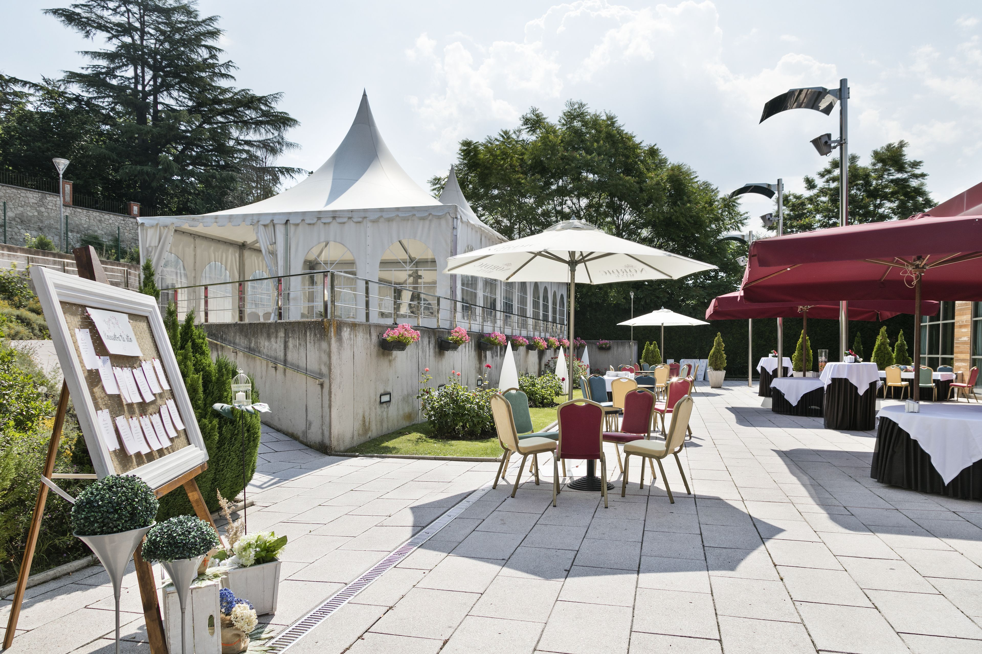 Een terras buiten met tafels en stoelen, een witte tent en potplanten.