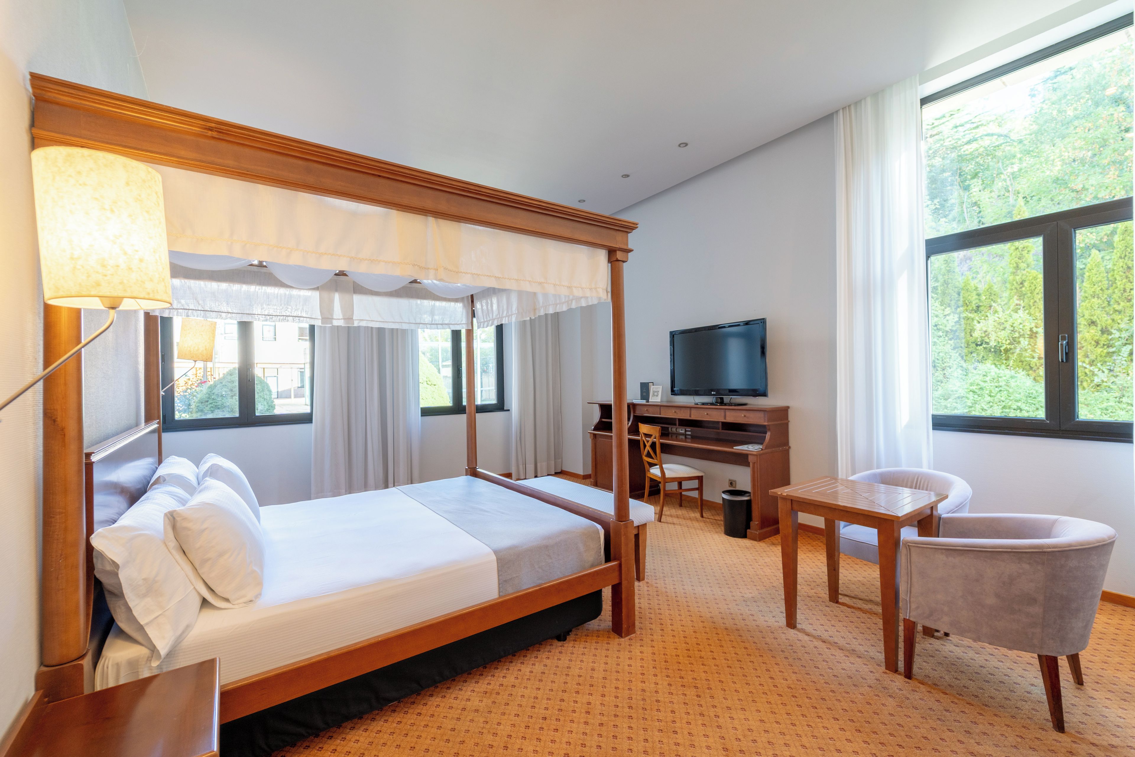 A hotel room with a four-poster bed, a desk with a chair, a television, and large windows.
