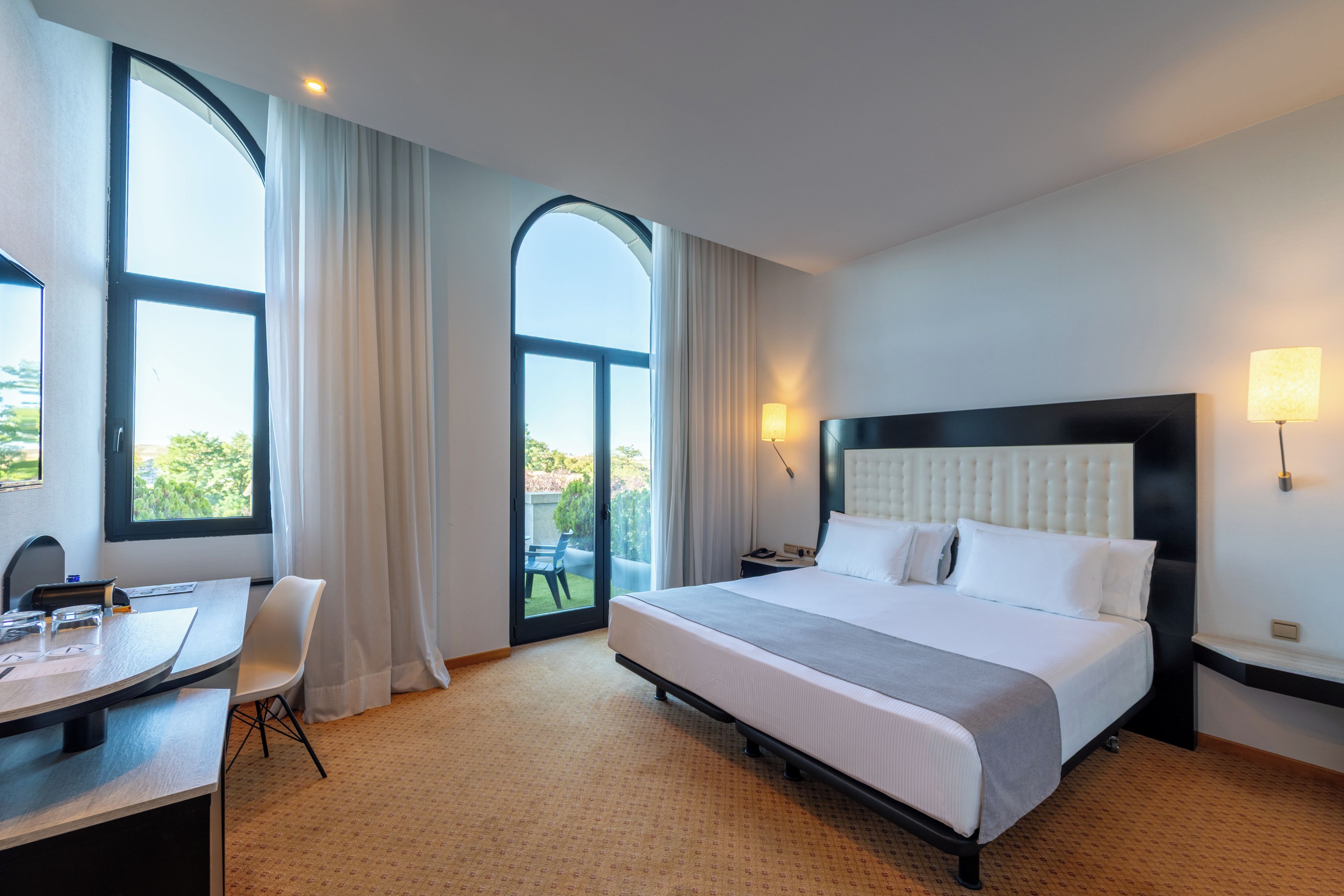 Ein Hotelzimmer mit einem Bett, einem Schreibtisch und einem Stuhl. Zwei Fenster und eine Tür führen zu einem Balkon.