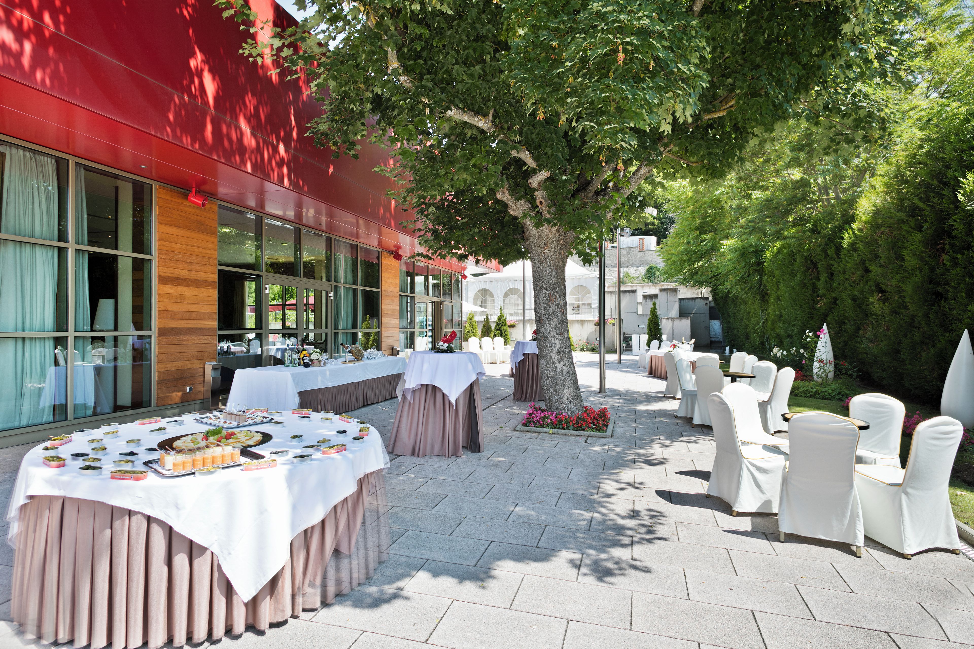 An outdoor dining area with tables set for a meal, surrounded by chairs and a tree.