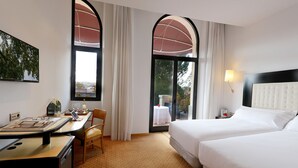 Ein Hotelzimmer mit zwei Betten, einem Schreibtisch mit einem Laptop, einem Stuhl und Blick ins Freie durch große Fenster.