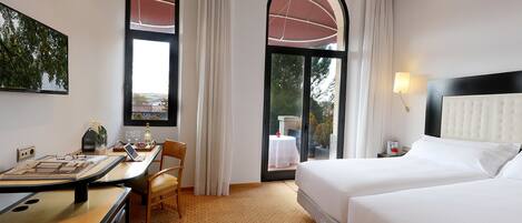 A hotel room with two beds, a desk with a laptop, a chair, and a view of the outdoors through large windows.