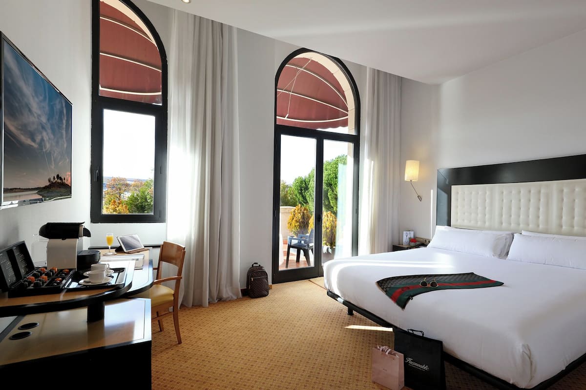 Habitació d'hotel amb llit, escriptori amb cafetera, cadira, finestra gran amb cortines i vista a l'exterior.