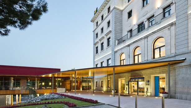 Ein Hotel mit einem modernen Glasvordereingang, einem Gebäude mit rotem Dach auf der linken Seite und einem klassizistischen Gebäude mit Rundbogenfenstern auf der rechten Seite.