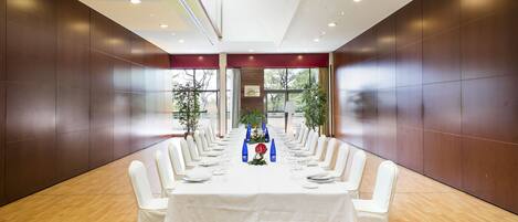 Un salón de conferencias con una larga mesa dispuesta para una reunión, rodeado de sillas blancas, y vistas a árboles a través de amplias ventanas.
