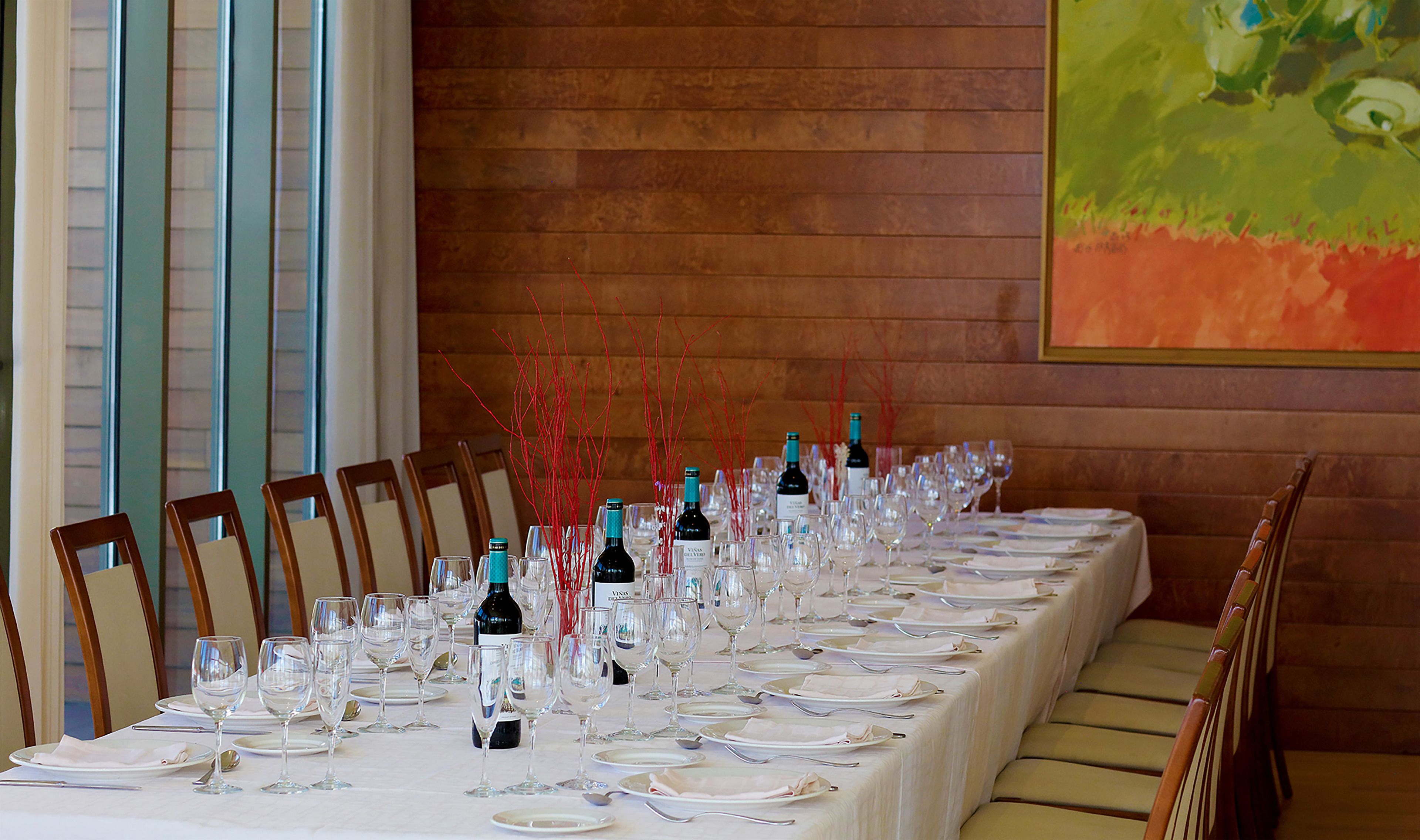 A long dining table set for a formal meal with wine bottles and glasses, wooden chairs, and a painting on the wall.