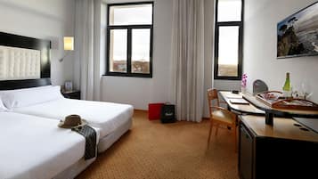 Quarto de hotel com duas camas, uma escrivaninha, uma cadeira, televisão e janela com vista.