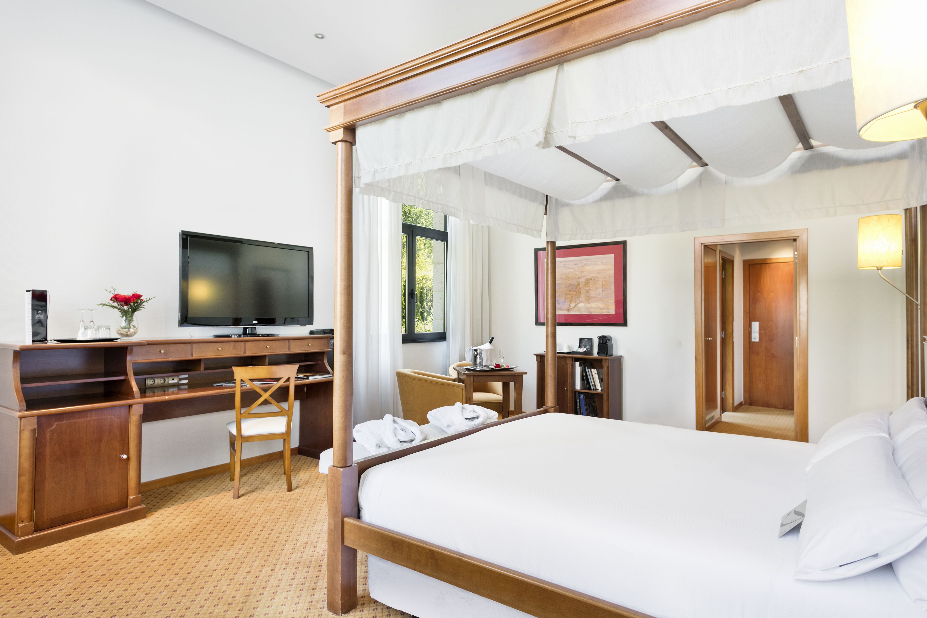 A spacious hotel room with a four-poster bed, a desk with a flat-screen TV, a chair, a small table, and a wardrobe.