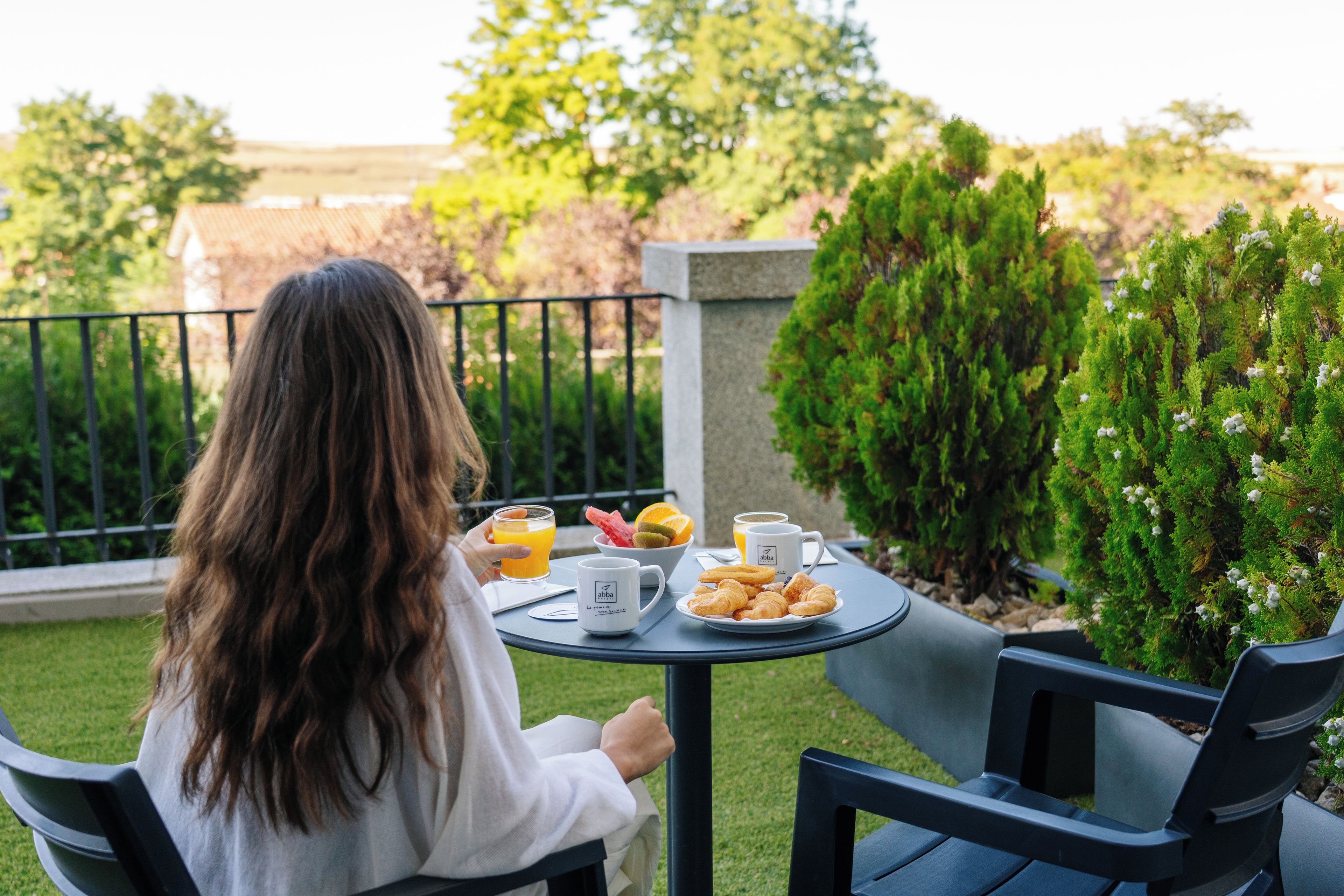 A person seated at a table with a meal, outdoors with greenery in the background.