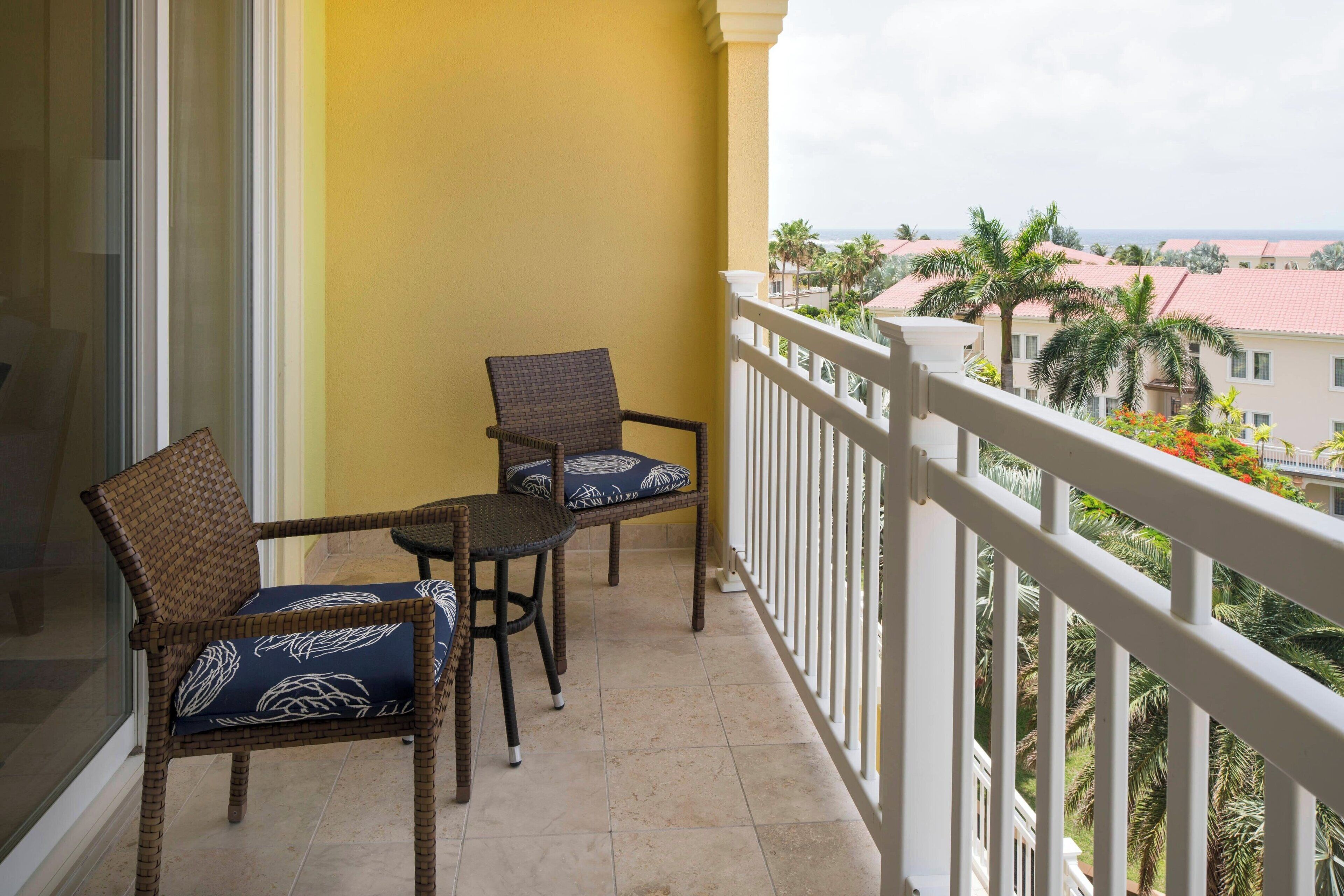 A balcony with wicker chairs and a small table, overlooking a residential area with palm trees and houses.