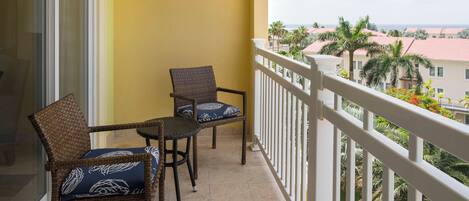 A balcony with wicker chairs and a small table, overlooking a residential area with palm trees and houses.