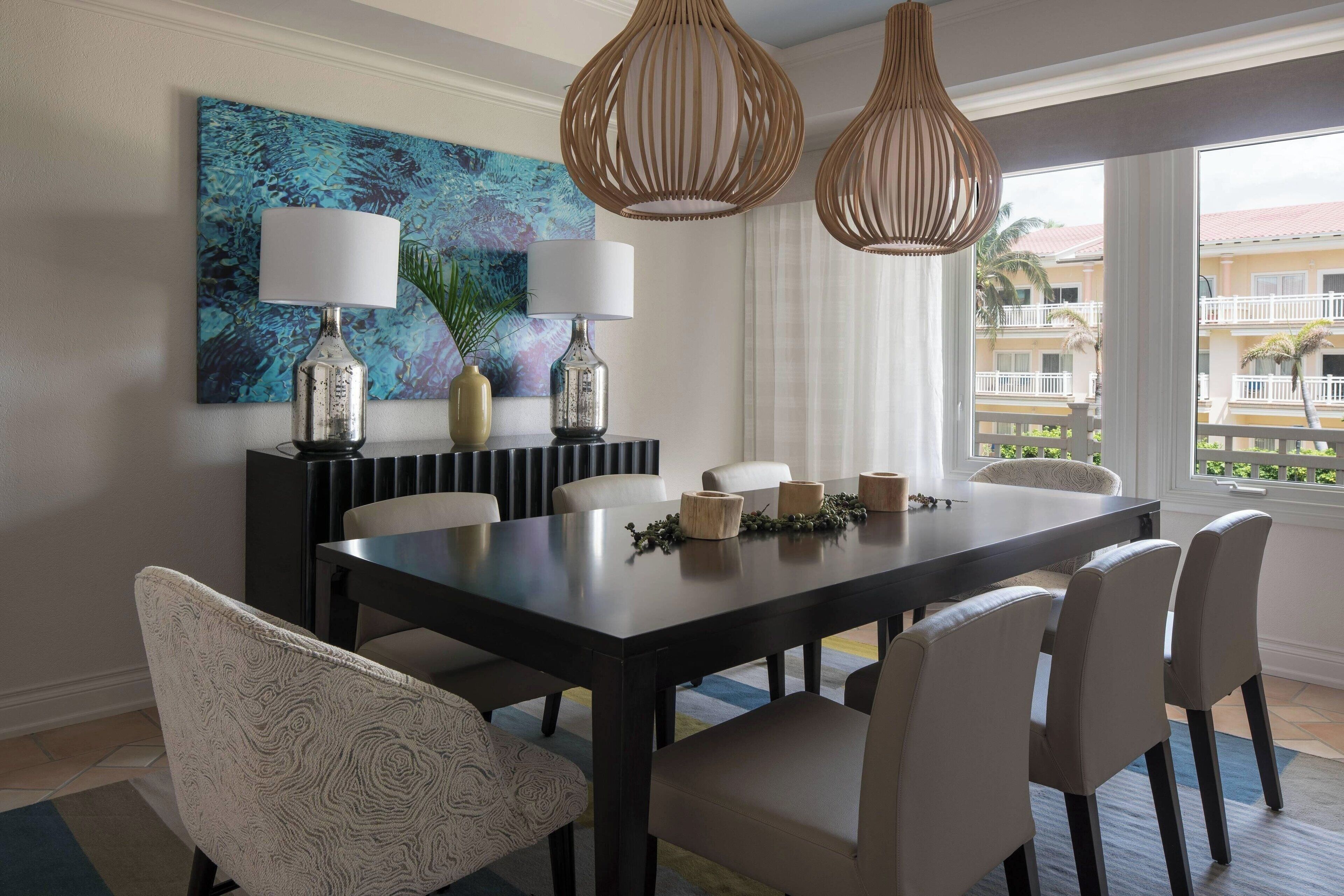 Uma área de refeições com uma mesa de madeira escura, cadeiras estofadas brancas e duas luminárias pendentes.
