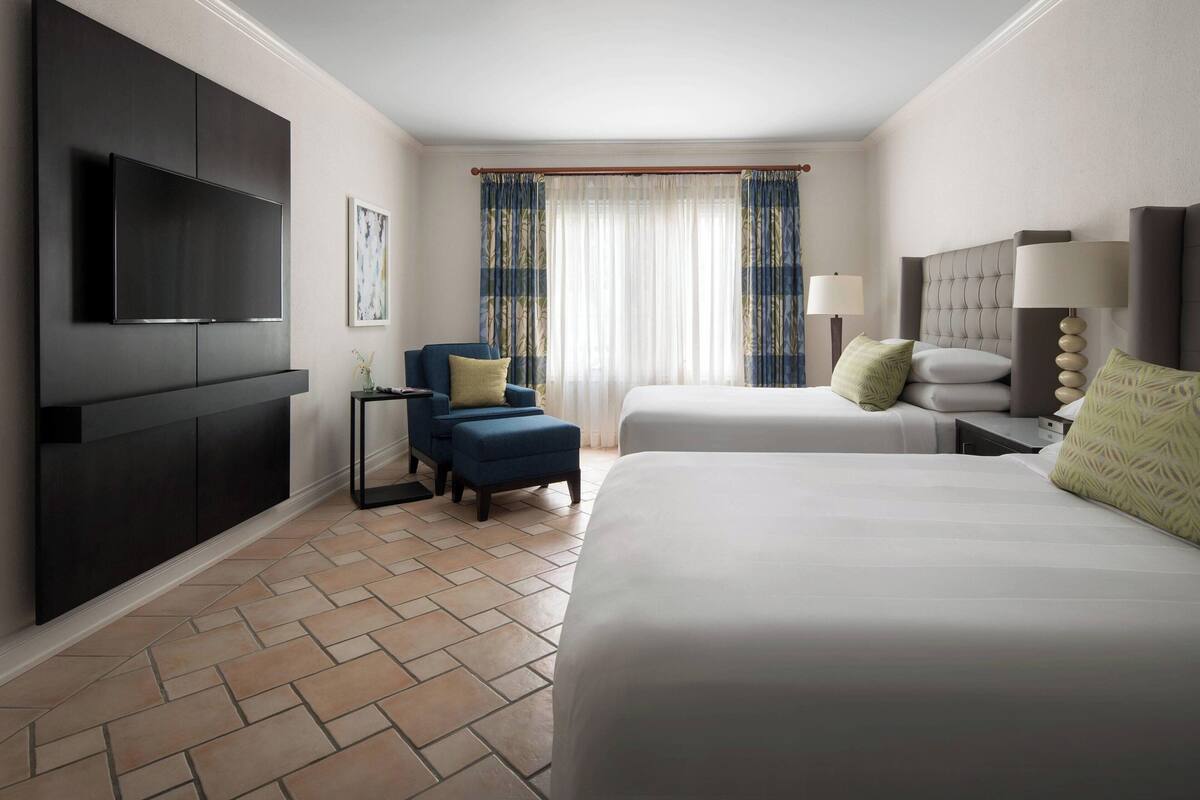 A hotel room with a large flat-screen TV, two beds, a chair, a nightstand, and a window with curtains.
