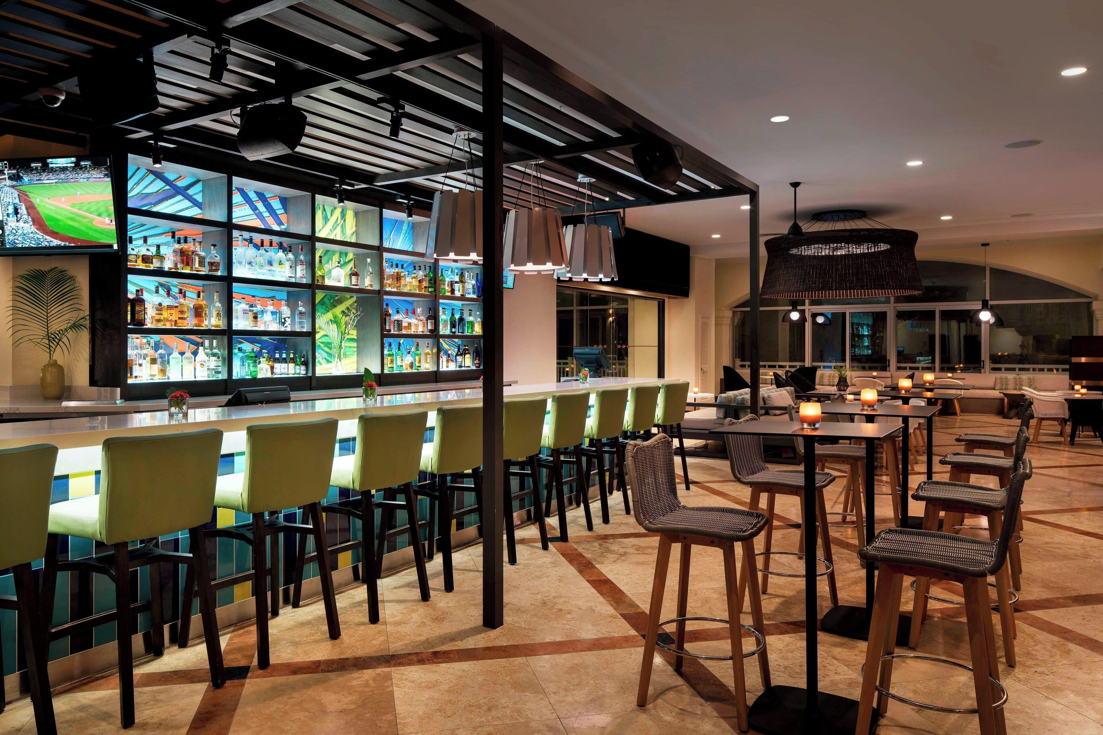 Een bar met diverse zitplaatsen, zoals barstoelen en gewone stoelen, en een selectie flessen op planken.