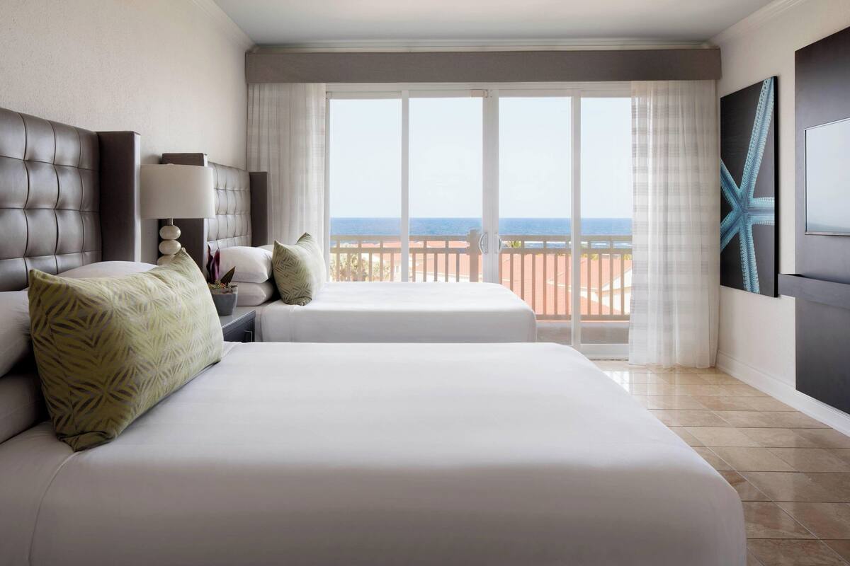 A hotel room with two beds, a nightstand, a lamp, a large window with a view of the sea, and a starfish artwork on the wall.