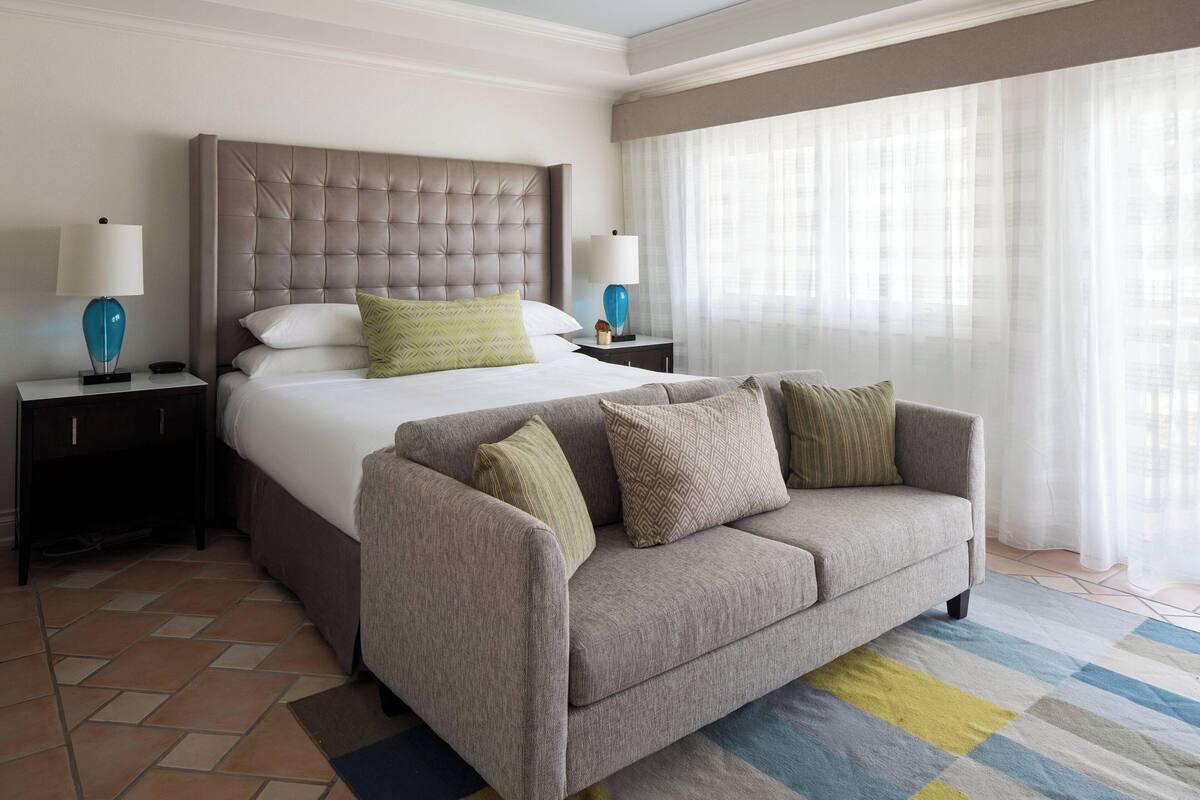 A hotel room with a large bed, a grey sofa, two bedside tables with lamps, and a window with sheer curtains.