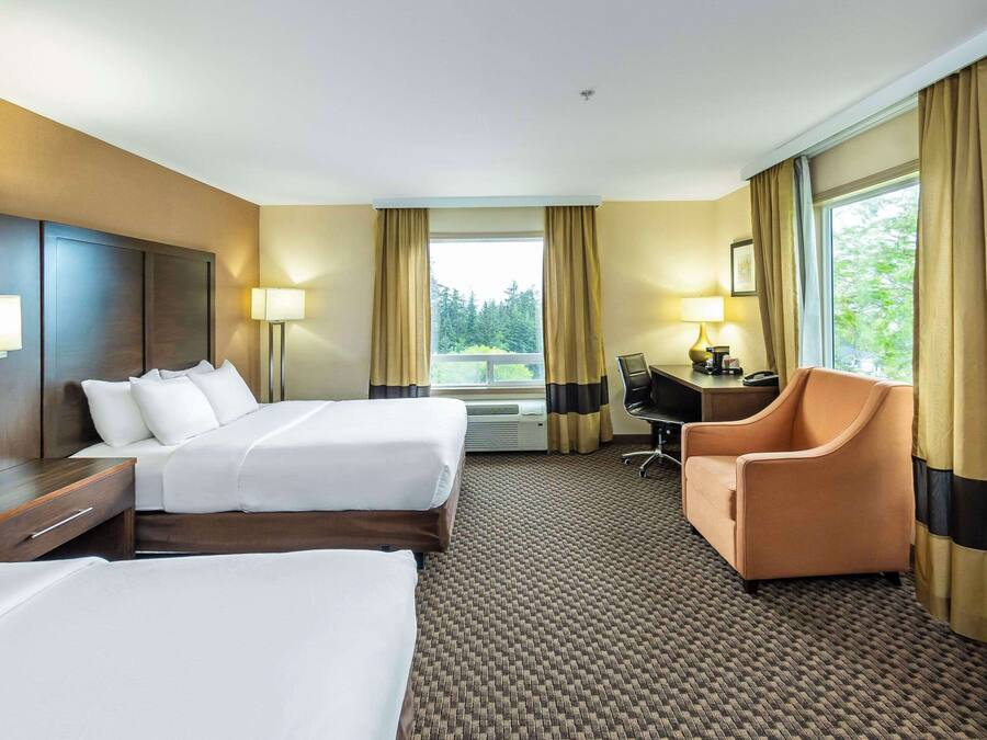Habitación de hotel con dos camas, un escritorio, una silla y un gran ventanal con cortinas.