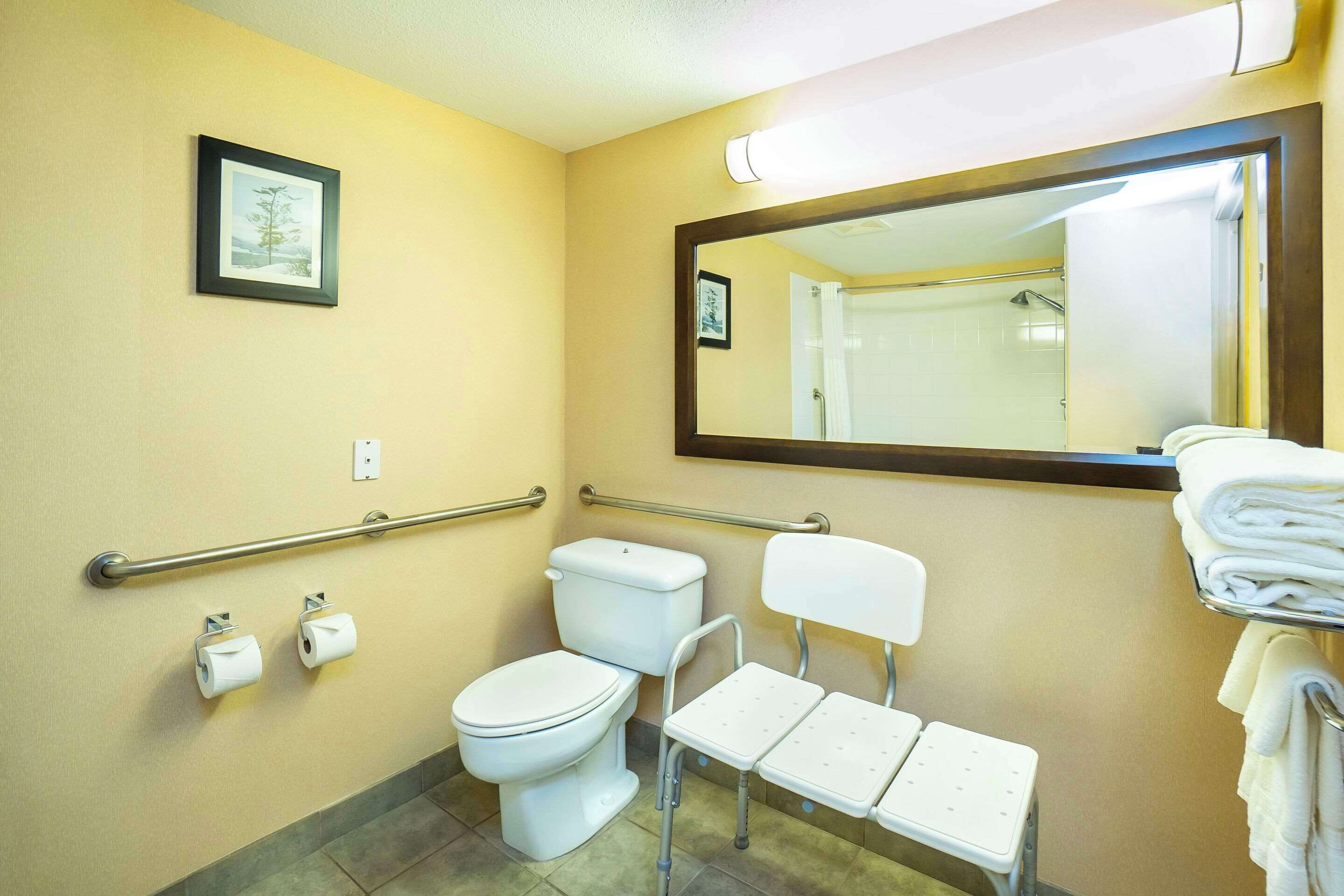 Un baño con inodoro, barras de apoyo, lavamanos, espejo y toallero.