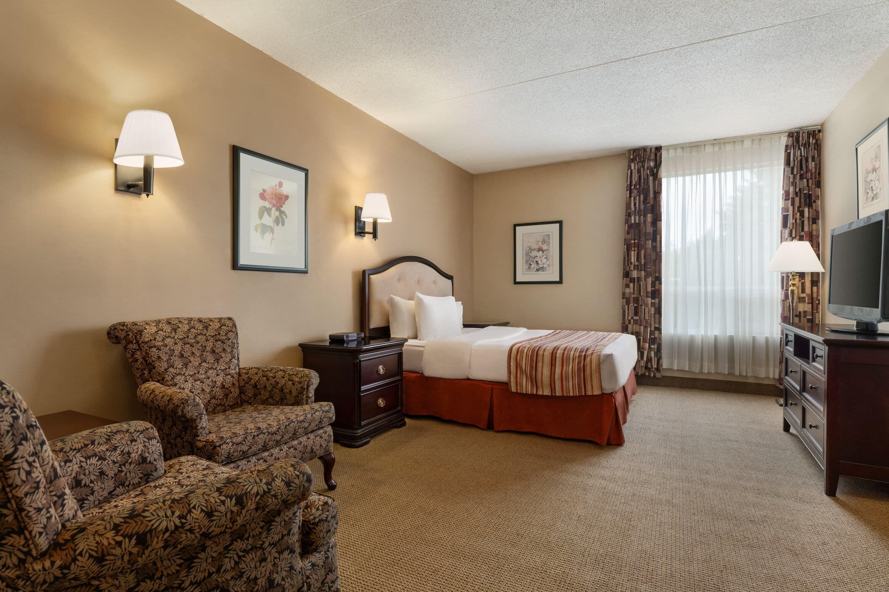 Quarto de hotel com cama, uma cadeira, mesa de cabeceira, televisão e duas luminárias de parede.