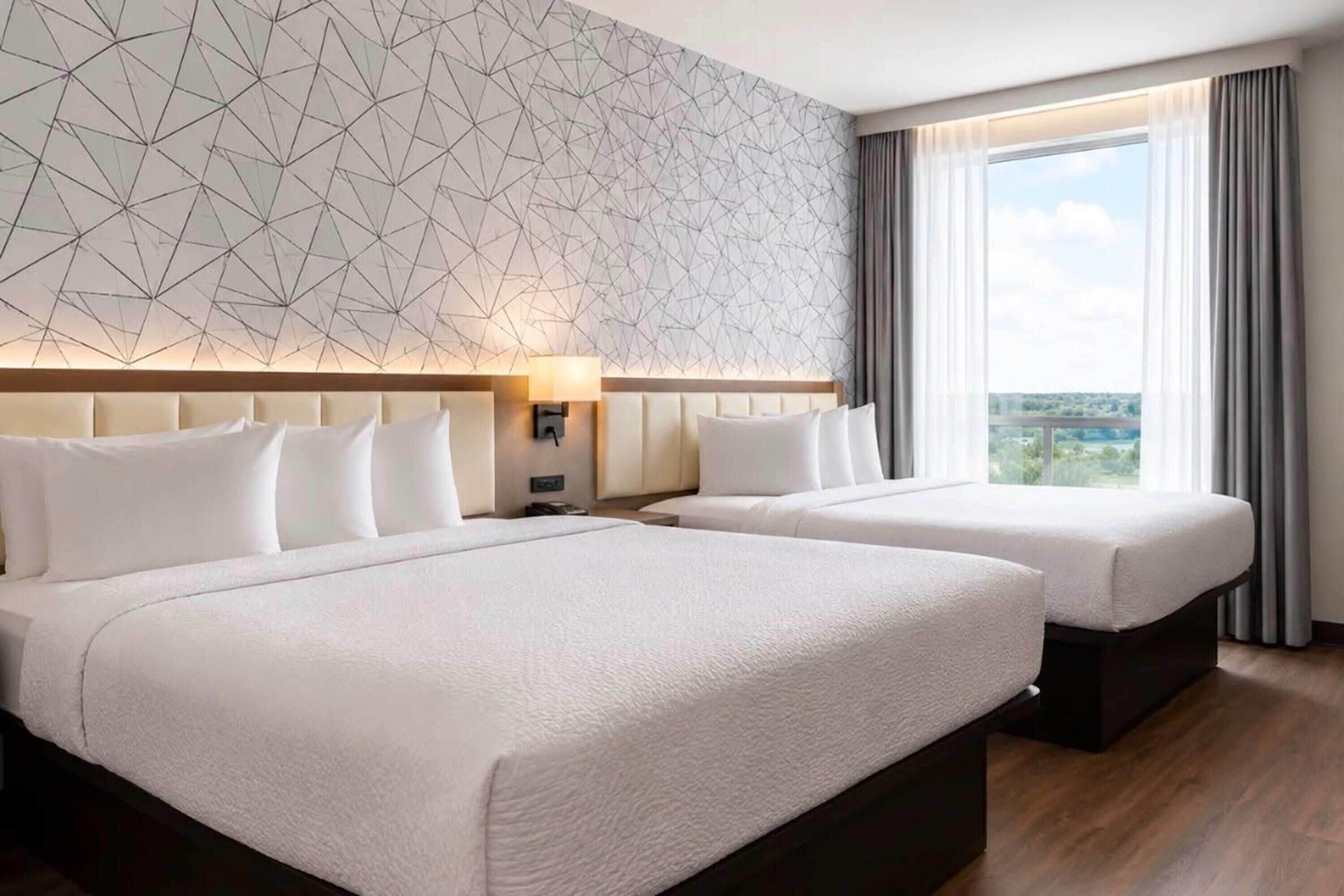 A hotel room with two beds, a geometric patterned wall, a large window with curtains, and a view of the outdoors.