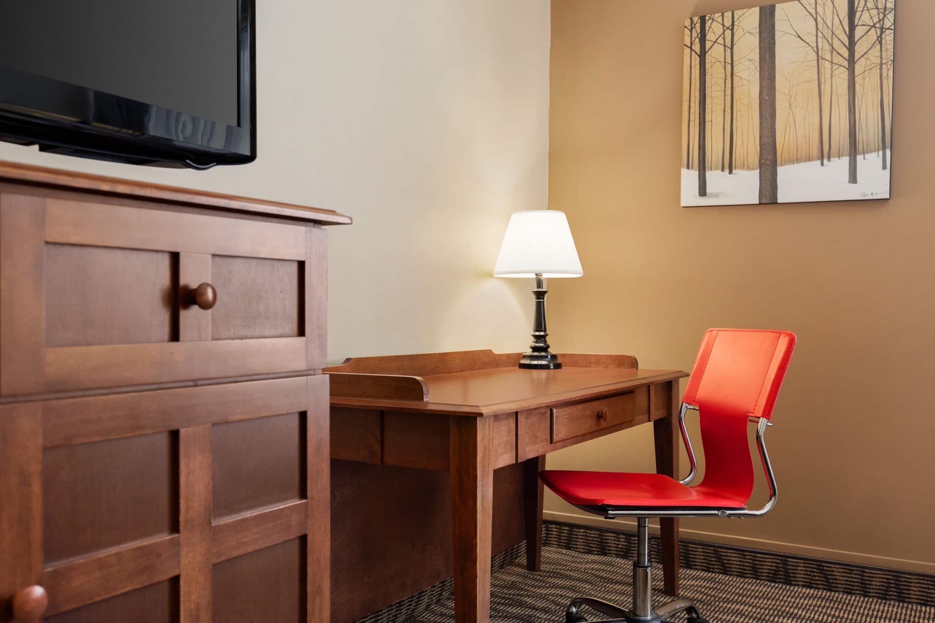 Une chambre d’hôtel avec un bureau en bois, une chaise rouge, une lampe, un téléviseur fixé au mur et un tableau représentant une forêt.