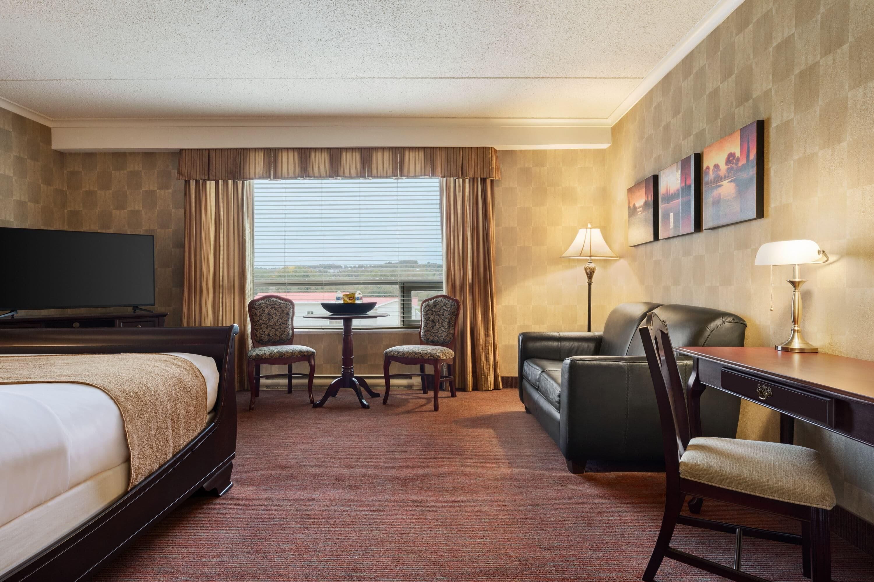 Habitación de hotel con cama, dos sillones, sofá, escritorio, televisor, mesa con un tazón y papas fritas, y ventana con cortinas.