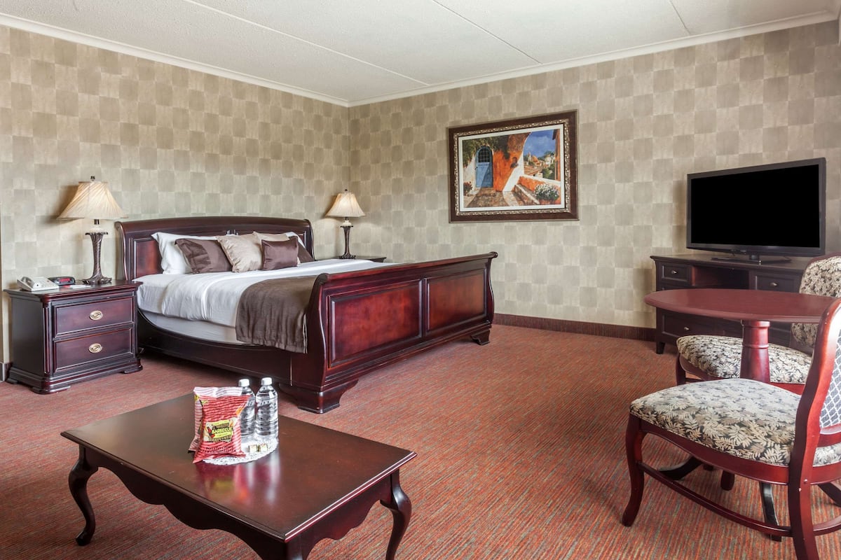 Habitación de hotel con una cama grande, dos mesitas de noche con lámparas, un televisor, una pequeña mesa con agua embotellada y aperitivos, y una silla con tapicería floral.