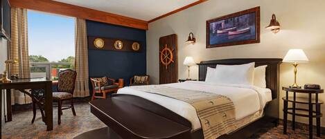 A hotel room with a large bed, a desk, two chairs, a ship's wheel decoration, a painting, and a view of the outdoors.
