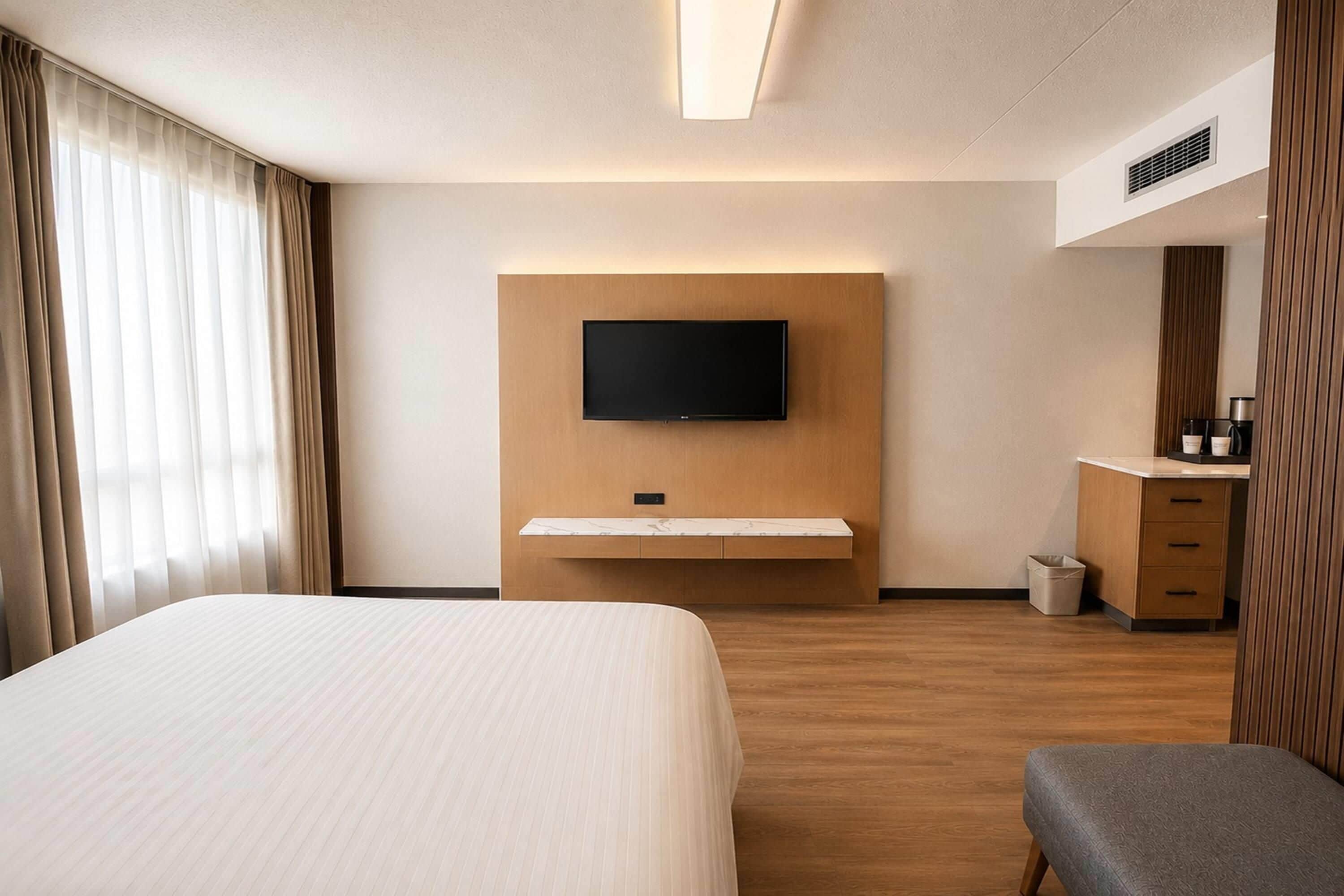 Habitación de hotel con cama, televisor de pantalla plana montado en un panel de madera, un escritorio pequeño con una silla y un cubo de basura.