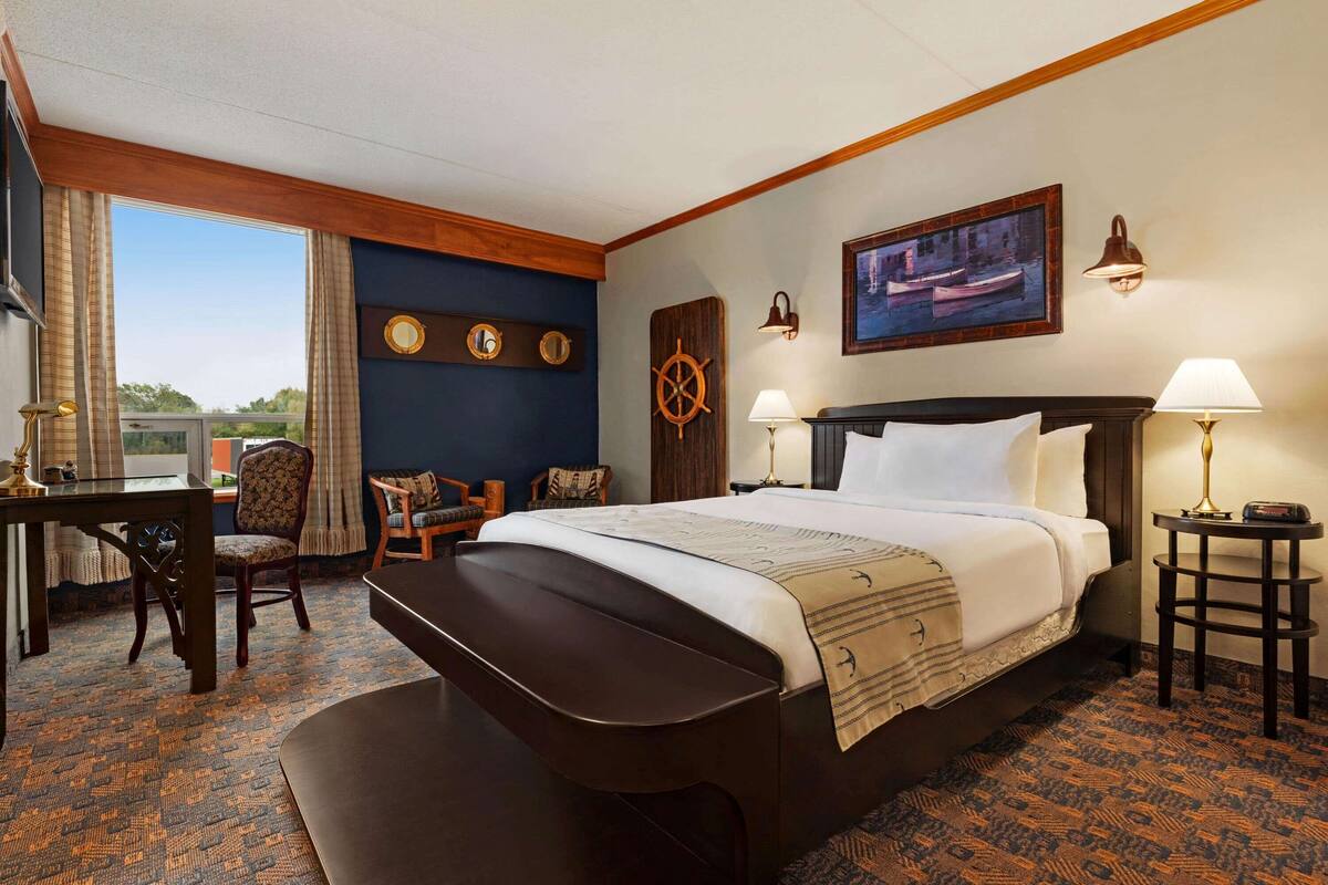 Habitación de hotel con una cama grande, un escritorio y una silla. Se ve una ventana con cortinas, un reloj de pared y un cuadro.