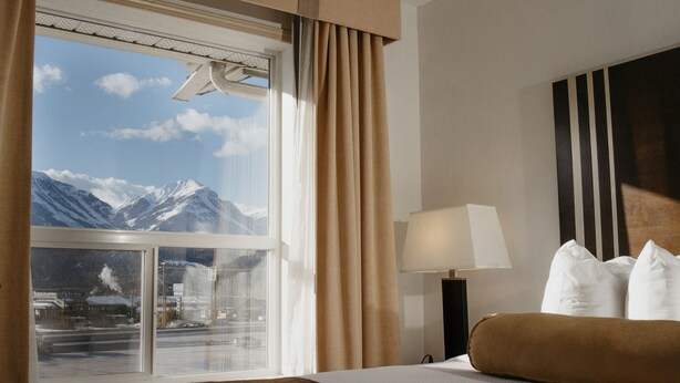 Habitación de hotel con un ventanal grande que ofrece vistas a la montaña, una cama con ropa de cama blanca y marrón, y una lámpara de noche.