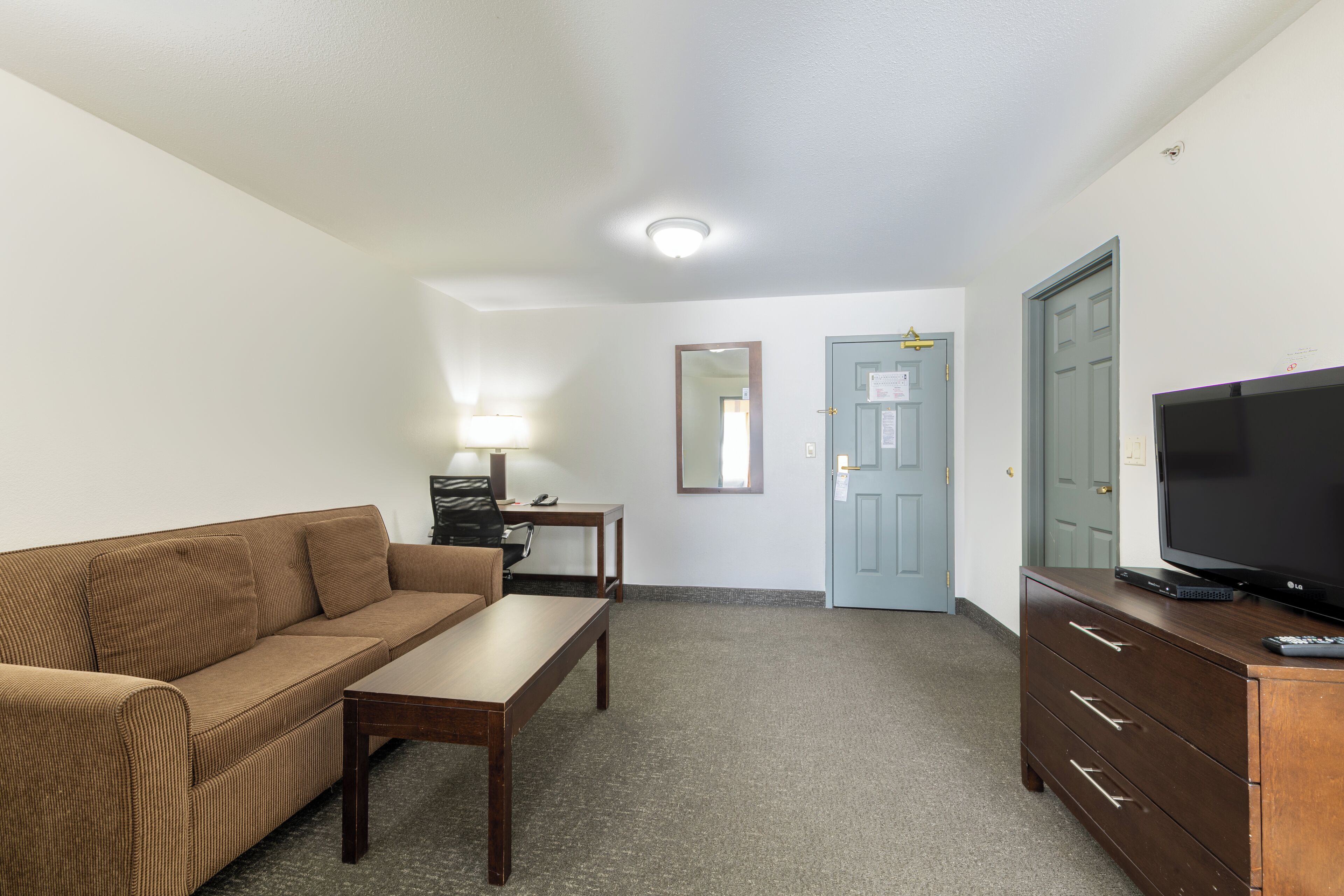 A compact living space with a sofa, a small table, a TV on a dresser, and a door with a light switch.