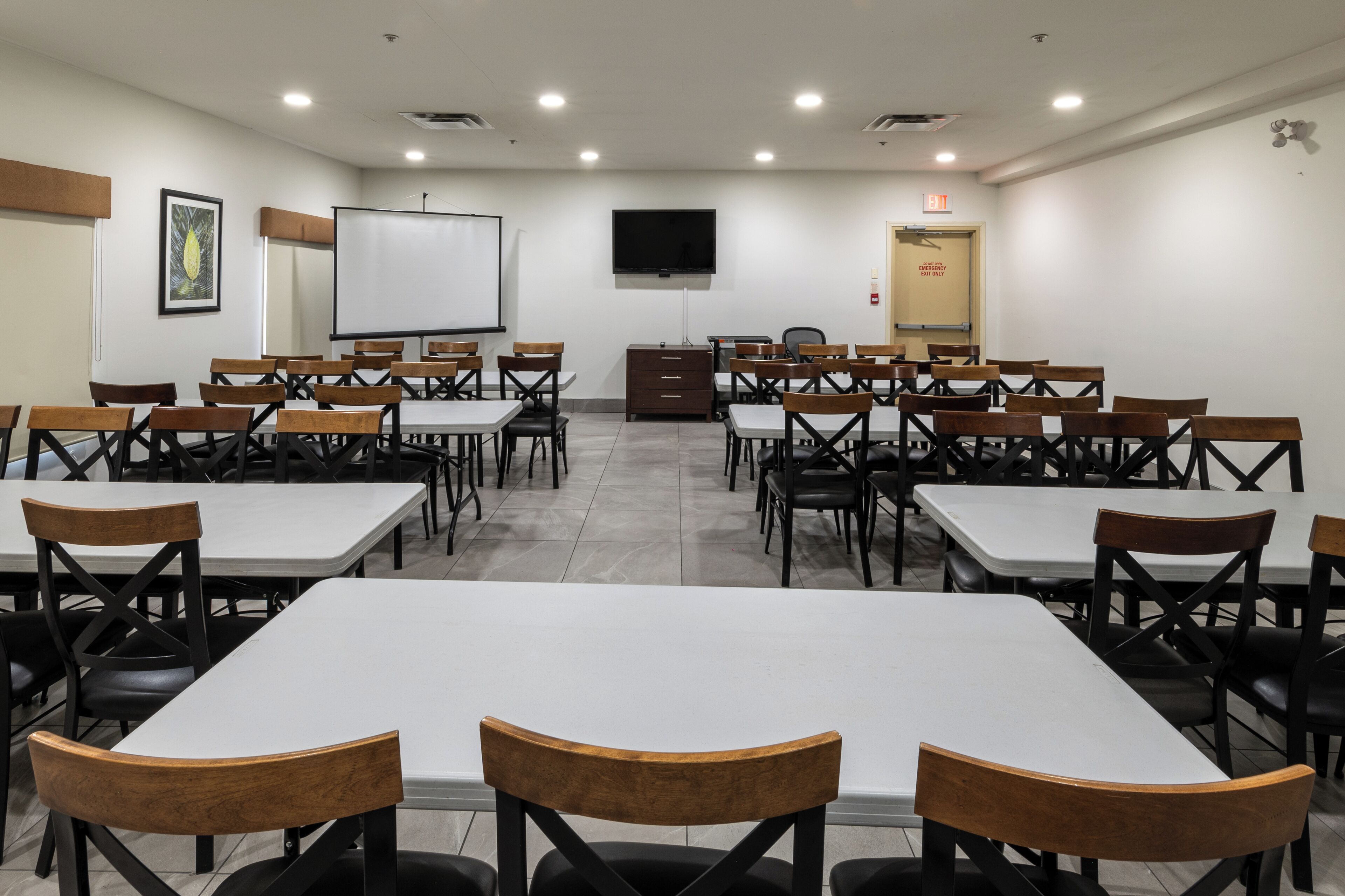 A conference room with white tables and chairs, a projector screen, a TV, and a door with an exit sign.