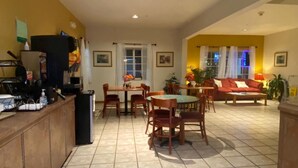 A dining area with a round table and chairs, a kitchen area with a refrigerator and counter, and a living area with a sofa and plants.