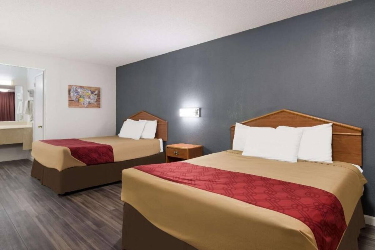 Dos camas dobles con cabeceros de madera, una mesita de noche de madera, una lámpara fijada a la pared y una vista parcial de un baño.