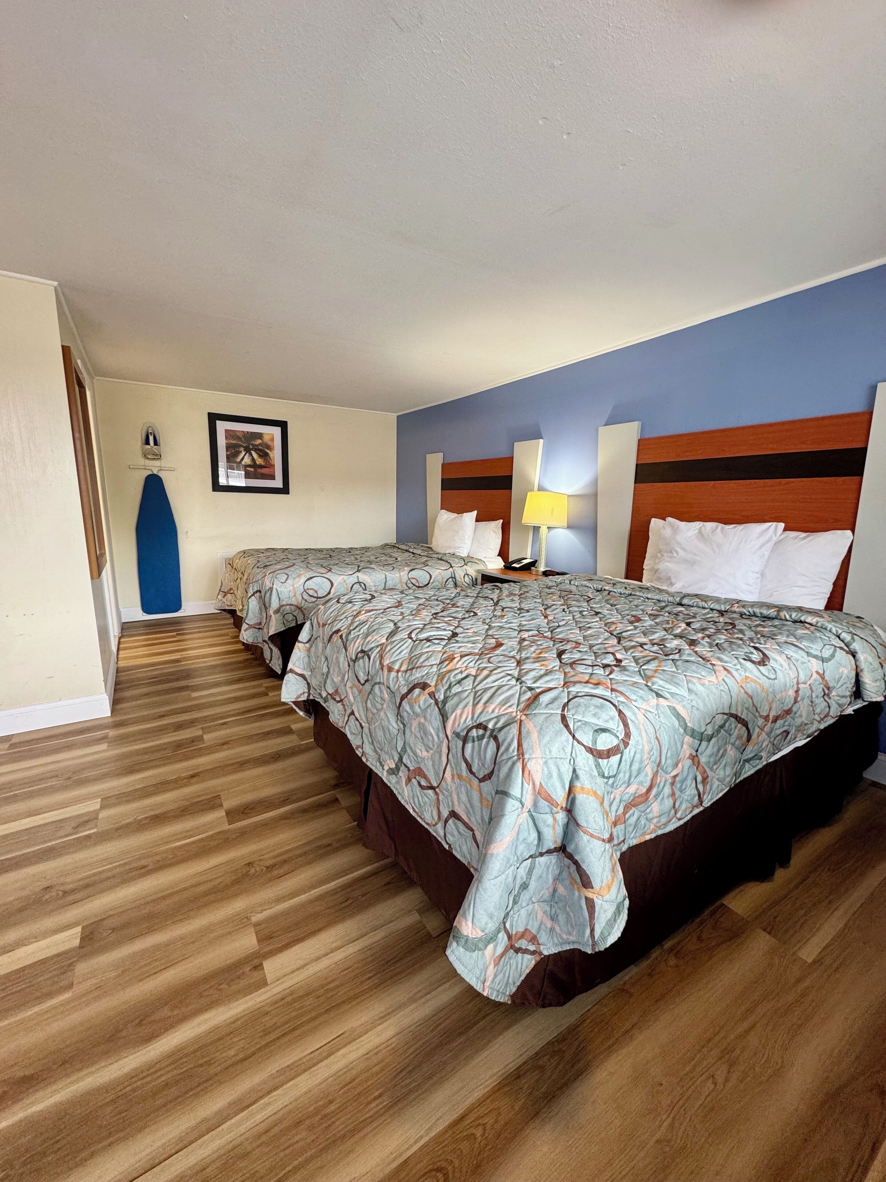 Una habitación de hotel con dos camas, suelo de madera, pared azul y un cuadro enmarcado.
