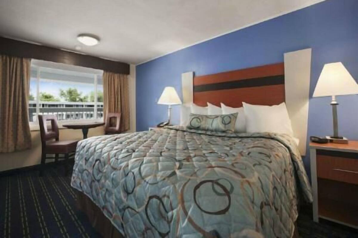 A hotel room with a bed, two bedside lamps, a nightstand, a chair, and a view of the outdoors.