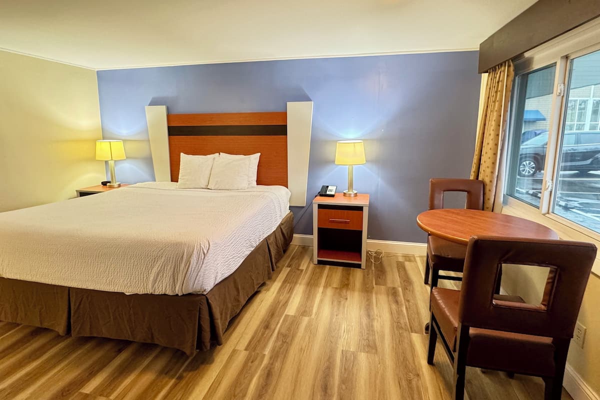 A hotel room with a large bed, two bedside tables with lamps, a wooden chair, and a window with a view of parked cars.