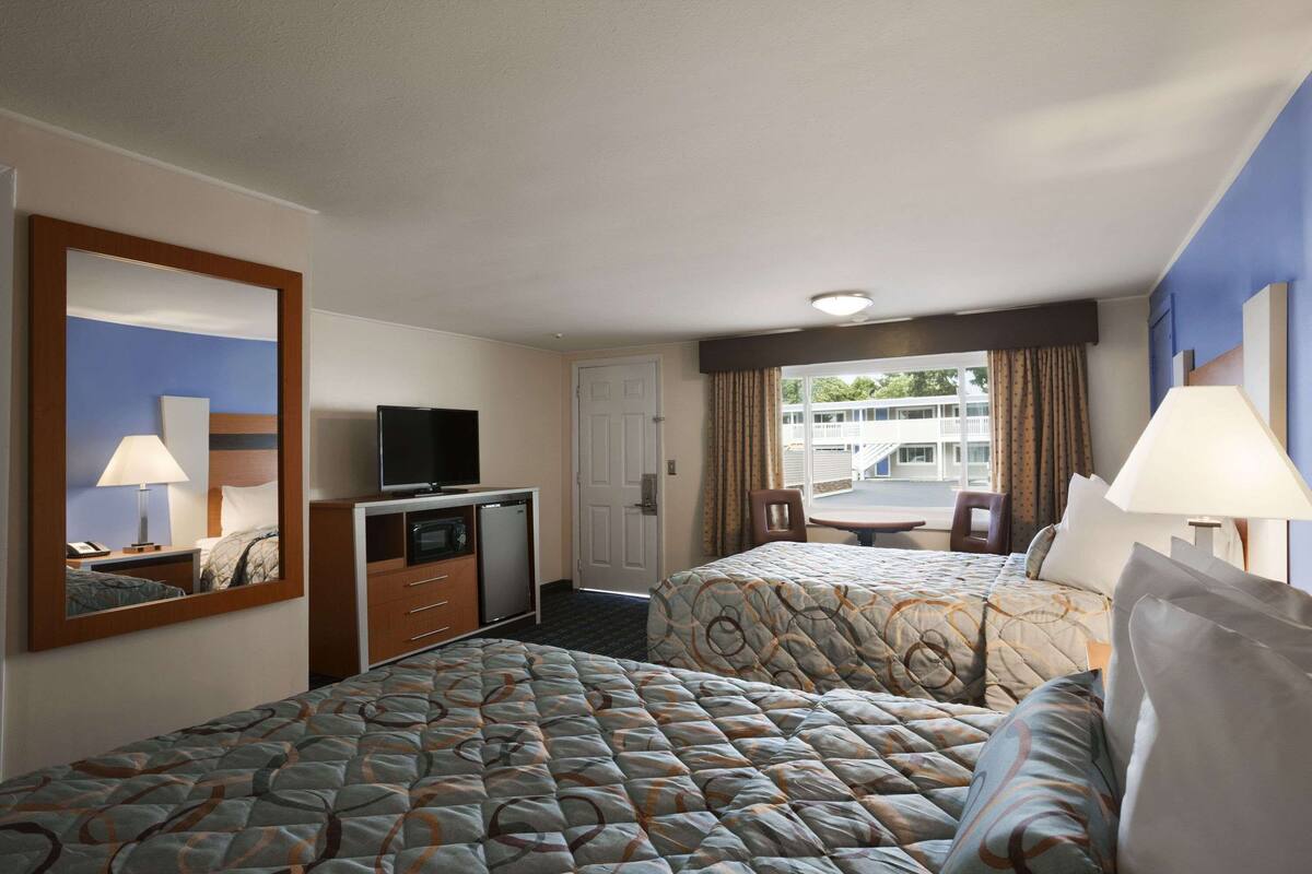 A hotel room with two beds, a desk with a television, a mirror, and a window with curtains.
