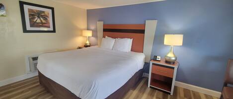 A neatly made bed with a wooden headboard, two bedside tables with lamps, a framed picture on the wall, and a wooden floor.