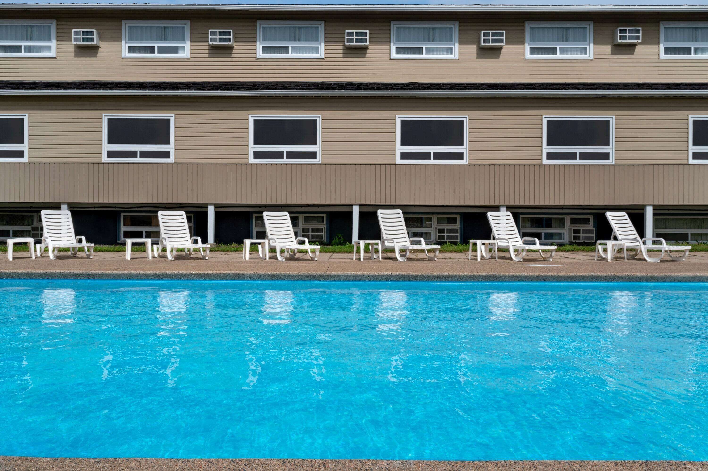 A swimming pool with white lounge chairs and a building with multiple windows and air conditioning units.