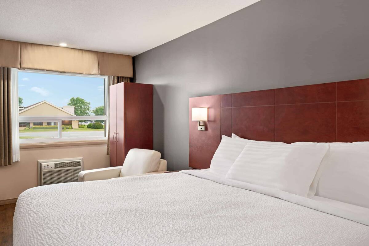 Une chambre d’hôtel avec un grand lit, une chaise, une fenêtre avec des rideaux et un luminaire fixé au mur.
