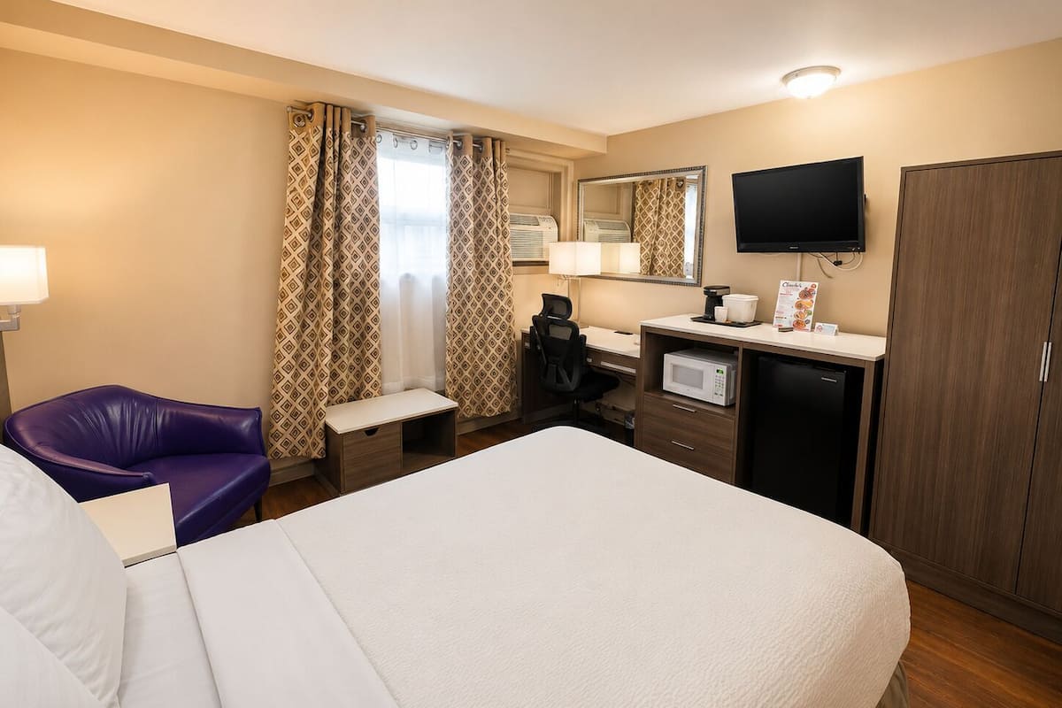 Une chambre d’hôtel équipée d’un lit, d’un bureau avec un ordinateur, d’un four à micro-ondes, d’une machine à café, d’une télévision et d’une armoire.