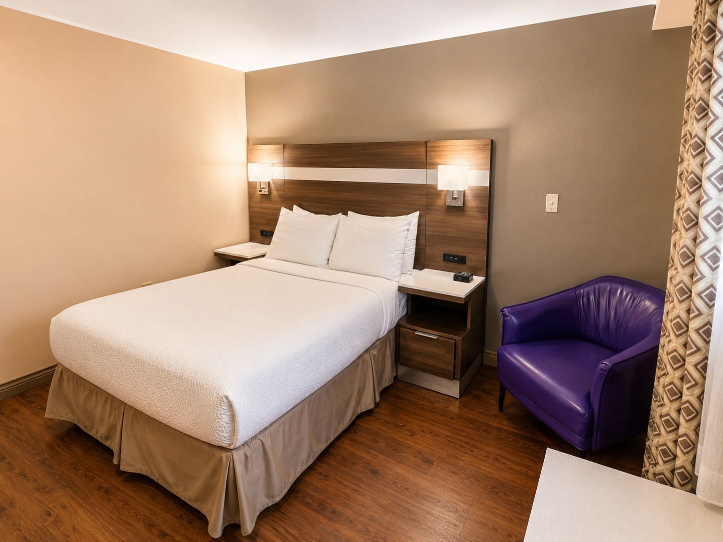 Una habitación de hotel con cama, mesitas de noche, un sillón morado y una cortina estampada.