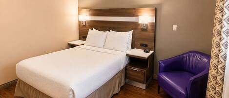 Una habitación de hotel con cama, mesitas de noche, un sillón morado y una cortina estampada.