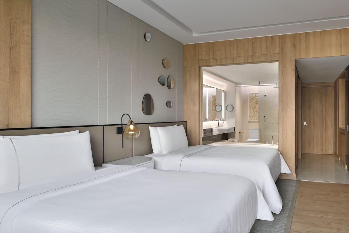 A hotel room with two beds, a wooden headboard, a wall-mounted mirror, and a bathroom visible through an open door.